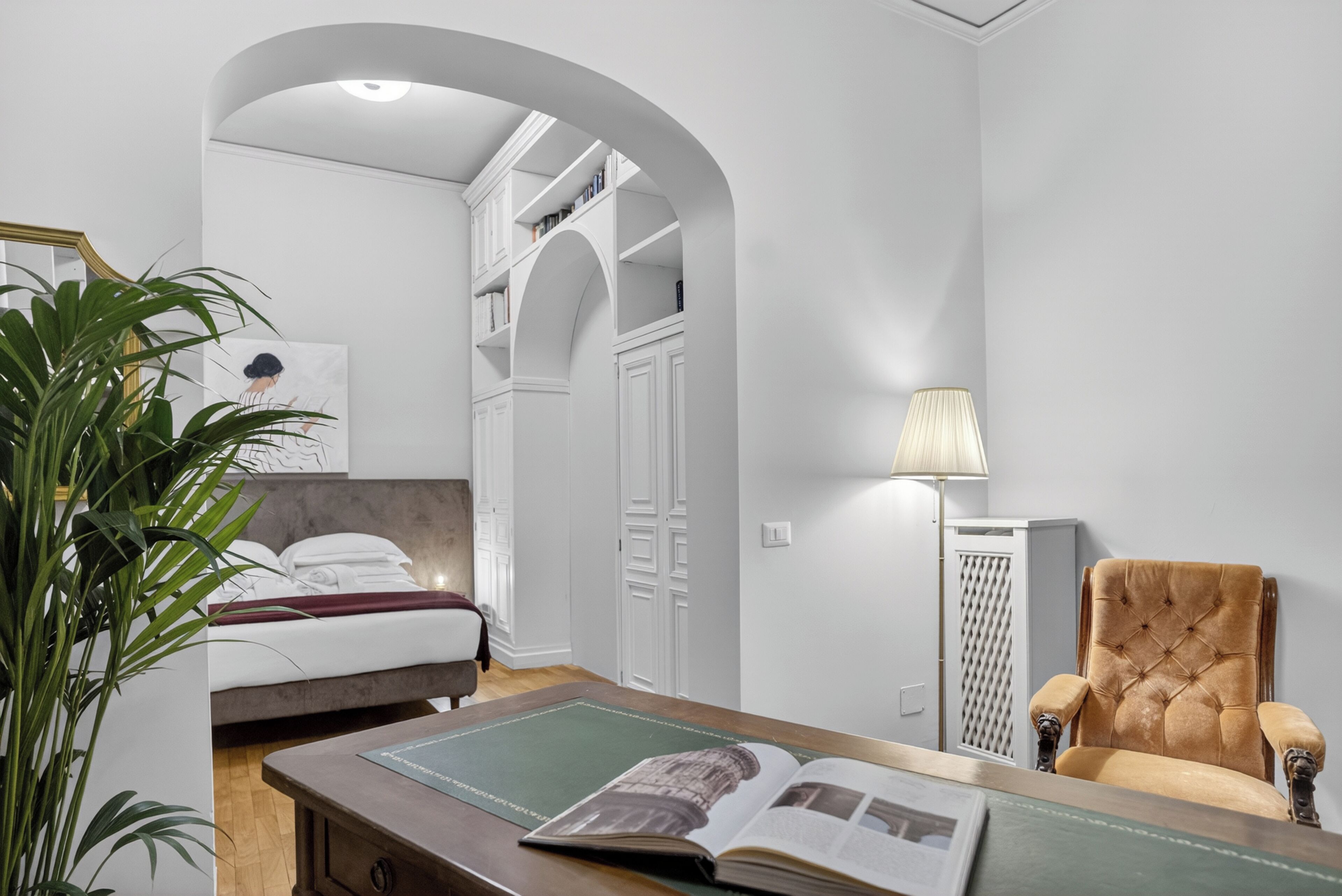 Un dormitorio con una cama, una zona de lectura con una silla y una mesa, una lámpara y una planta grande.