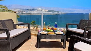 Une terrasse meublée de mobilier en osier, une table avec des aliments pour le petit-déjeuner et une vue sur la mer et les montagnes lointaines.