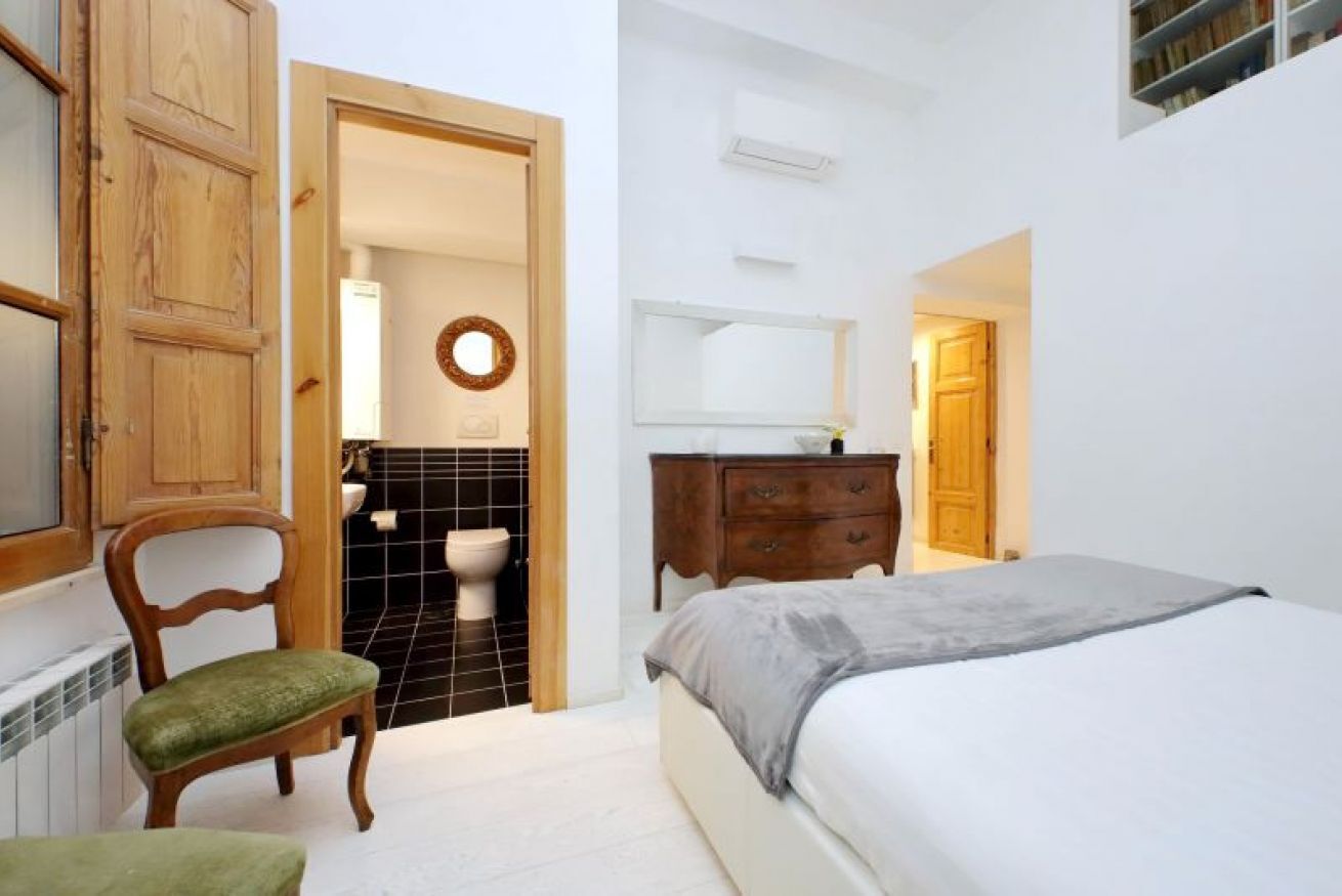 Un dormitorio con cama, una silla de madera, un aparador, un espejo y un baño visible a través de una puerta abierta.