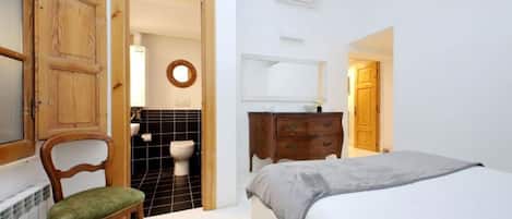 Un dormitorio con cama, una silla de madera, un aparador, un espejo y un baño visible a través de una puerta abierta.