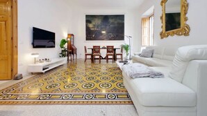 Una sala de estar con un sofá blanco, una mesa de comedor con sillas, un televisor montado en la pared y un cuadro grande sobre la mesa.