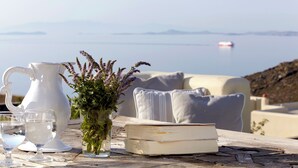 Une table avec une carafe, des verres, un livre et un vase de fleurs, le tout placé devant un paysage côtier.