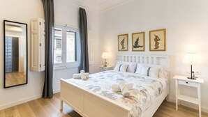 Ein Schlafzimmer mit einem Bett, einem Nachttisch, einer Lampe, einem Fenster mit Vorhängen und Wandbildern.