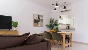 Ett modernt vardagsrum med en soffa, ett matbord med stolar, en tv och ett köksområde med skåp och en spegel.