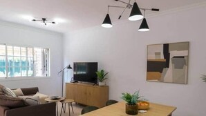 Ett modernt vardagsrum med en soffa, ett matbord med stolar, en tv placerad på ett träskåp och en taklampa.