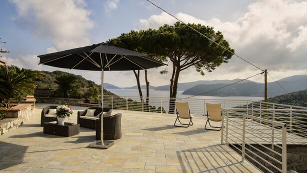Een terras buiten met rieten meubels, een grote parasol en een prachtig uitzicht op zee en bergen.