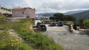 Een roze villa met een verharde oprit en geparkeerde auto's, met uitzicht op een waterlichaam en bergen.