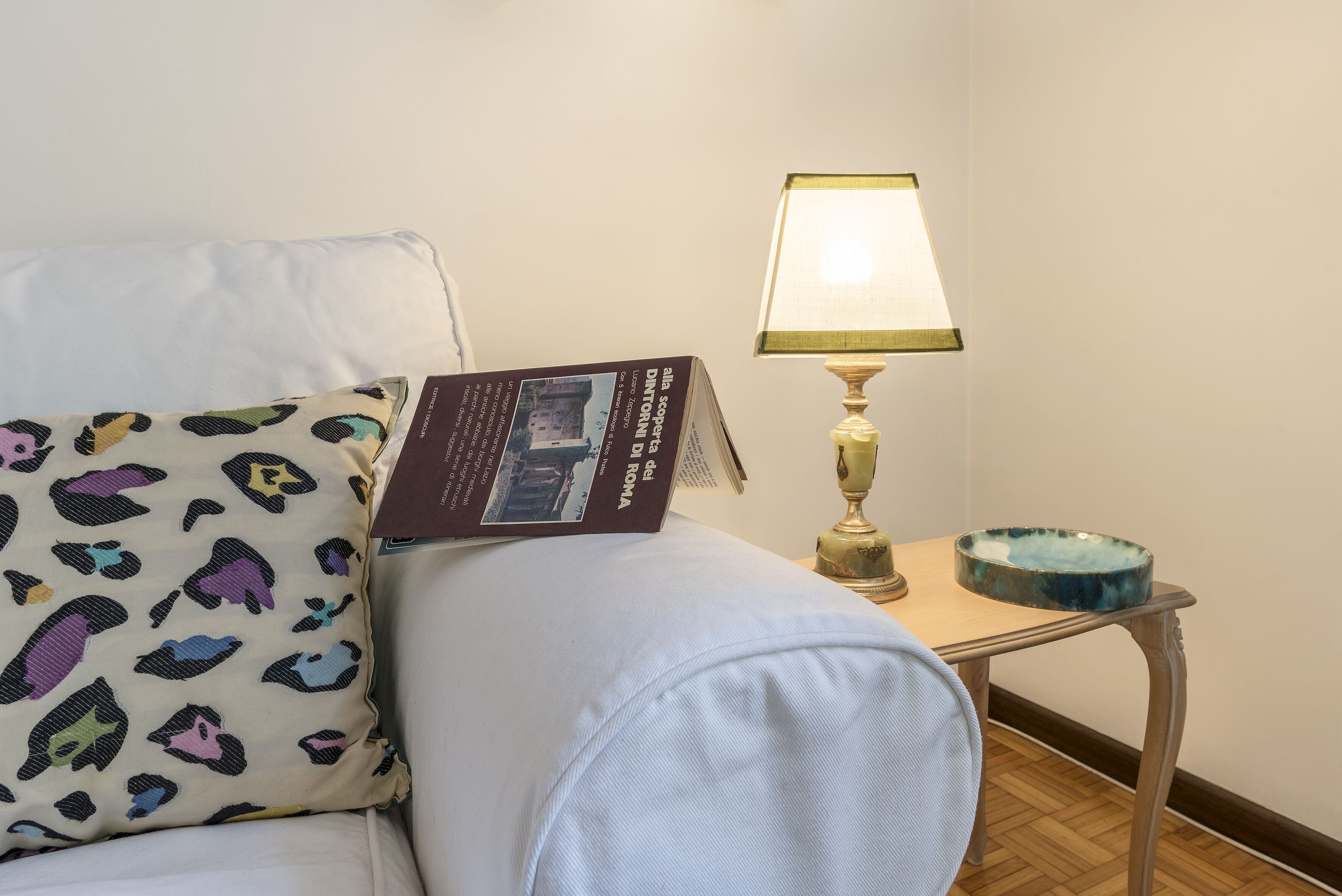A room with a bed, a reading lamp, a small table with a bowl, and a pillow with a pattern.