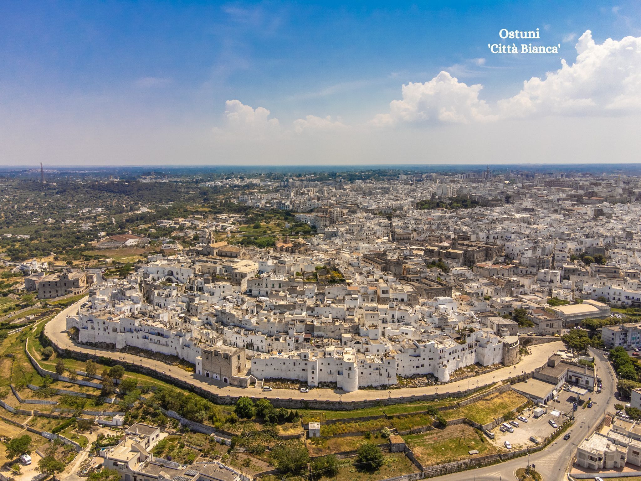 Vista aérea de Ostuni, 'Città Bianca', con una zona destacada de muros blancos, carreteras circundantes y un paisaje urbano distante.