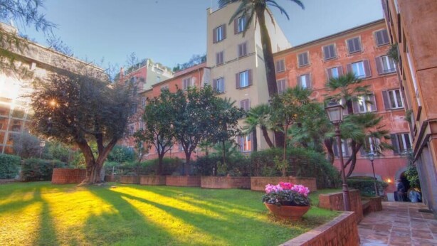 A courtyard with a palm tree, a flower bed, and a building with multiple windows and balconies. - BEAUTIFUL ROMAN APARTMENT NEXT TO TREVI FOUNTAIN (Roma)