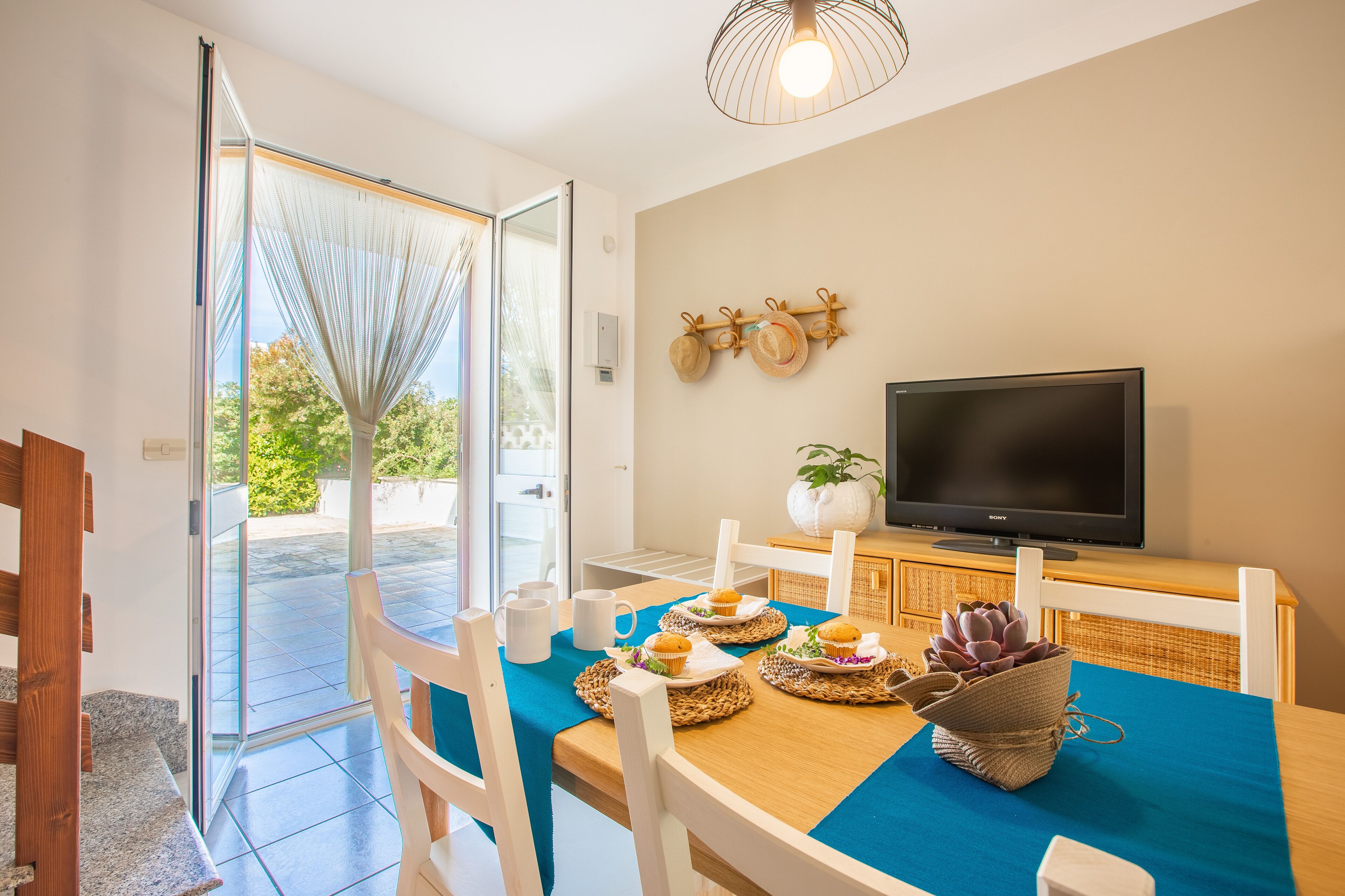 A dining area with a table set for four, a television on a stand, and a view of the outdoors through a glass door.