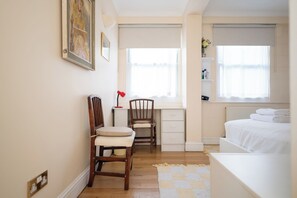 A bedroom with a bed, two wooden chairs, a desk with a lamp, a radiator, and a window with blinds. - Brilliantly Located 2BD Flat by Buckingham Palace (London)