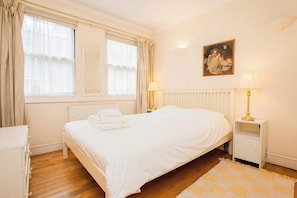 A bedroom with a bed, bedside tables, a radiator, a window with curtains, and a painting on the wall. - Brilliantly Located 2BD Flat by Buckingham Palace (London)