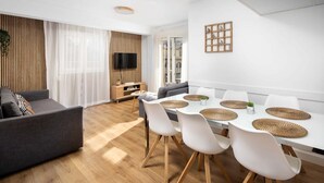 Ein modernes Wohnzimmer mit grauem Sofa, einem Esstisch mit weißen Stühlen und einem Fernseher auf einem Holzschrank.