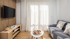 Ein modernes Wohnzimmer mit einem Flachbildfernseher an einer Wand aus Holzpaneelen, einem grauen Sofa mit gemusterten Kissen, einem kleinen weißen Tisch mit dekorativer Schale und transparenten Vorhängen, die natürliches Licht hereinlassen.
