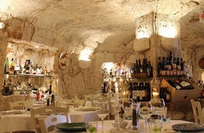 Un ristorante con tavoli apparecchiati, bottiglie di vino sugli scaffali e interni che ricordano una grotta di pietra.