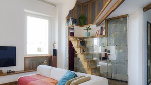 Uma sala de estar moderna com um sofá branco, um cobertor vermelho e laranja, e uma escada de madeira que leva a uma porta de vidro.