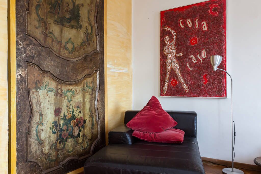 A room with a vintage door, a red abstract painting, a black sofa with a red pillow, and a floor lamp.