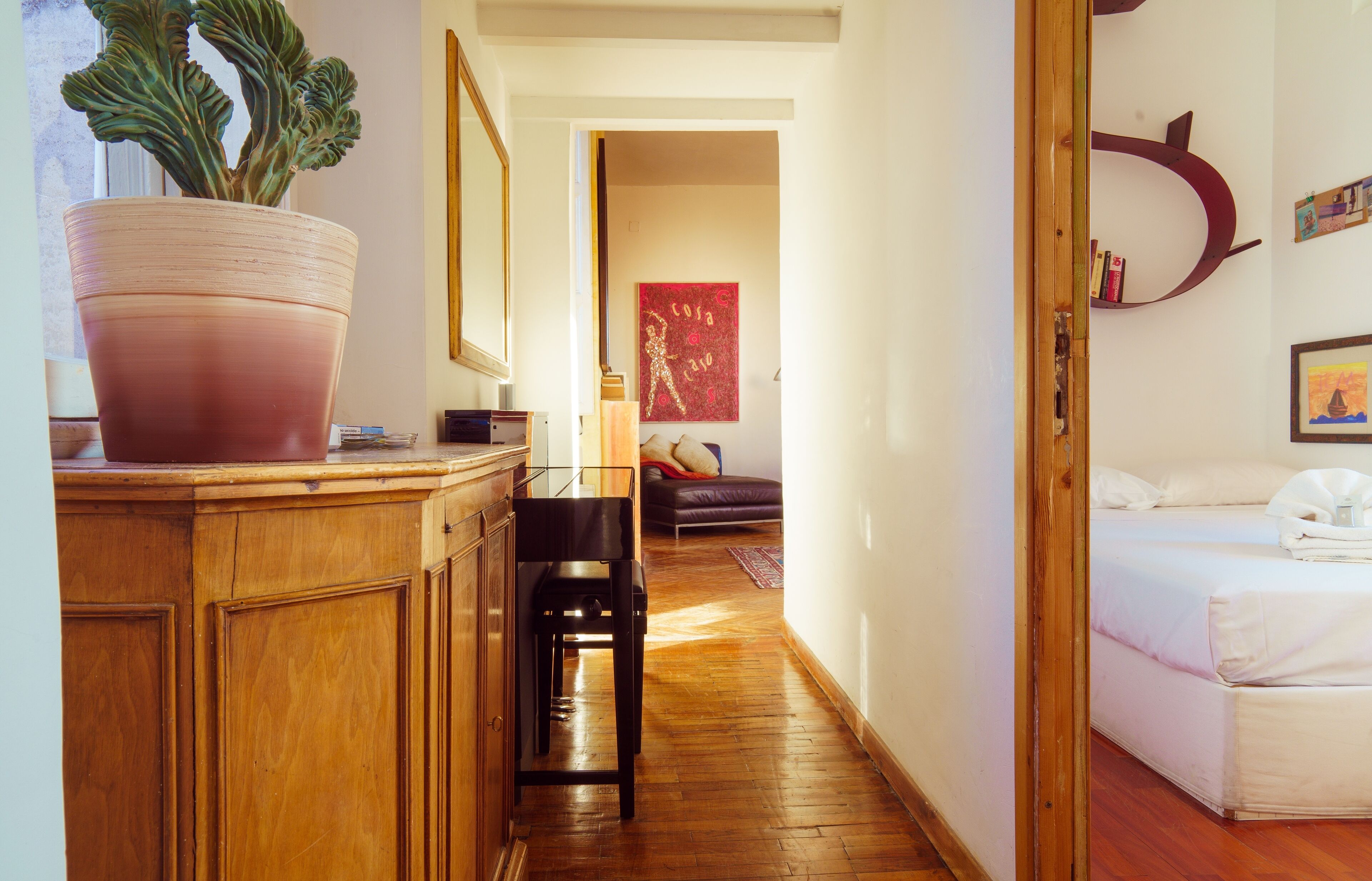 Une chambre d’hôtel avec un plancher en bois, une grande plante en pot, un lit, un bureau avec une chaise et une bibliothèque.