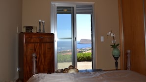 Ein Schlafzimmer mit einem Holzkasten, einem Bett mit weißer Bettdecke, einem Topf mit einer Orchidee und Blick auf den Ozean durch eine Glastür.