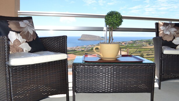 Ein Balkon mit Korbmöbeln, einem Tisch mit Tasse und Untertasse sowie Blick auf das Meer und die Landschaft.