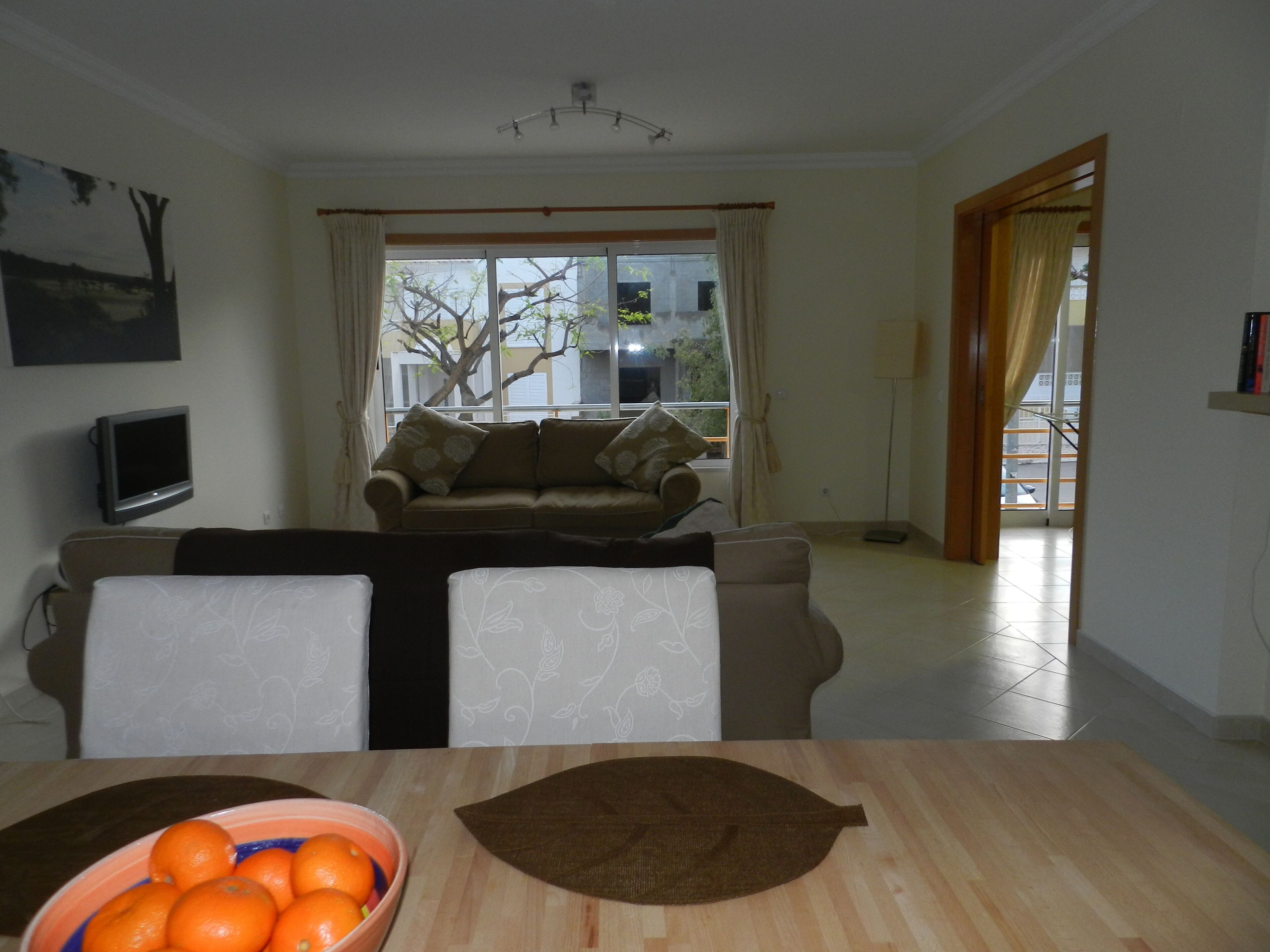 Ein Wohnzimmer mit einer Couch, einem Esstisch mit einer Schüssel Orangen, einem Fernseher und einem Fenster mit Vorhängen.