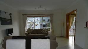 A living room with a sofa, a dining table with a bowl of oranges, a TV, and a window with curtains.