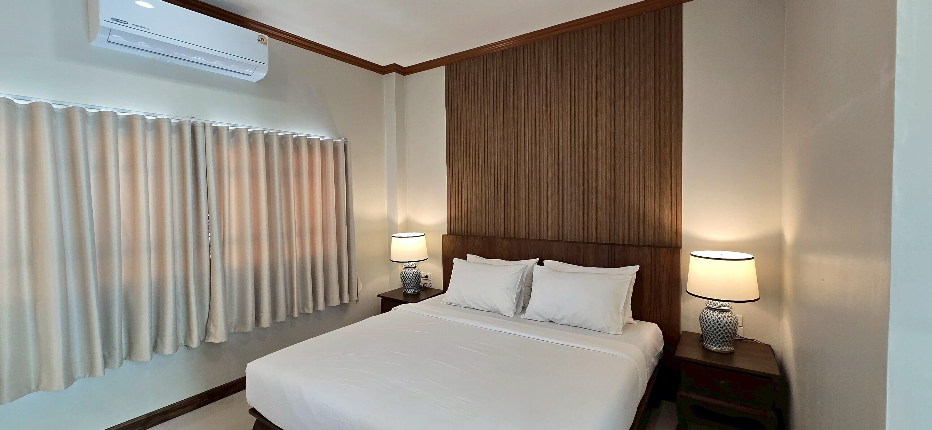 Habitación de hotel con una cama grande, dos lámparas de noche, un cabecero de madera, un aire acondicionado instalado en la pared y una ventana con cortinas.