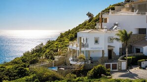 A white building with a pool, surrounded by greenery and overlooking a body of water.
