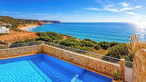 A swimming pool with a stone wall, overlooking a beach and ocean.