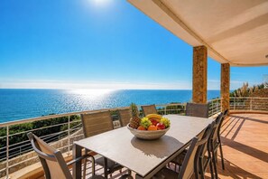 A balcony with a table, chairs, and a bowl of fruit, overlooking a coastal view.