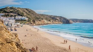 A coastal beach with buildings on a hillside, clear blue water, and people enjoying the beach.
