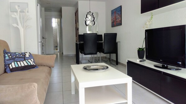 A modern living room with a sofa, a coffee table, a TV, and a dining area with chairs and a table.