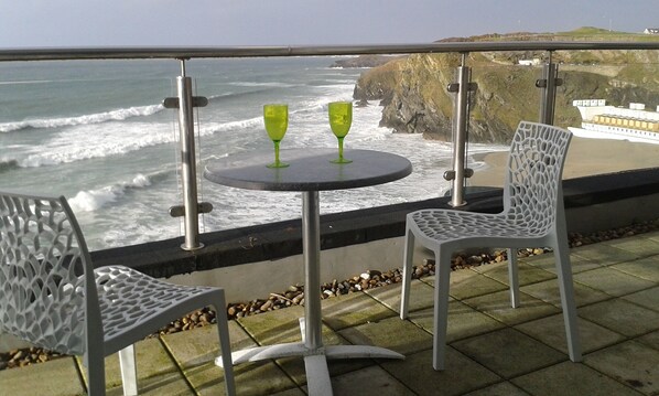 A balcony with two chairs and a table, overlooking a rocky coastline with waves crashing against the shore. - Rocklands studio with incredible views across the sea and beaches and cliffs (newquay. cornwall)