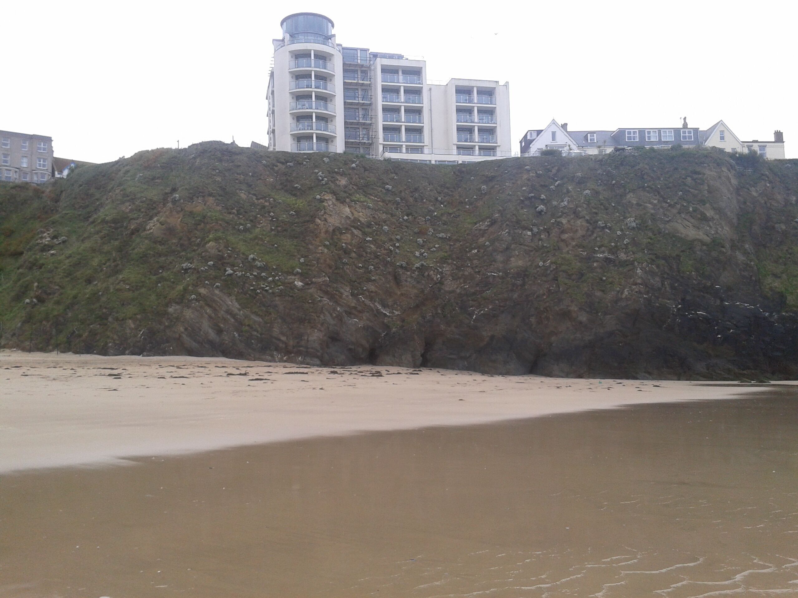 A coastal area with a sandy beach, a rocky cliff, and buildings in the background.
