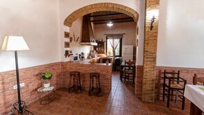 A kitchen with a central island, a dining area with chairs, a small table with a lamp, and a potted plant.