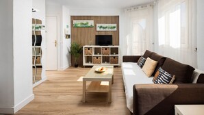 Ein modernes Wohnzimmer mit einer Couch, einem Couchtisch und einem an einer Holzwand montierten Fernseher. Auf den Regalen befinden sich dekorative Körbe und ein Blumentopf.