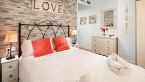 Ein Schlafzimmer mit Steinwand, einem Bett mit weißer Bettwäsche und orangefarbenen Kissen, einem Nachttisch mit Lampe, einer Kommode und Wandbildern.