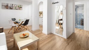 Ein modernes Wohnzimmer mit einem Glastisch, ein Essbereich mit weißem Tisch und Stühlen sowie eine durch einen Bogen sichtbare Küche.