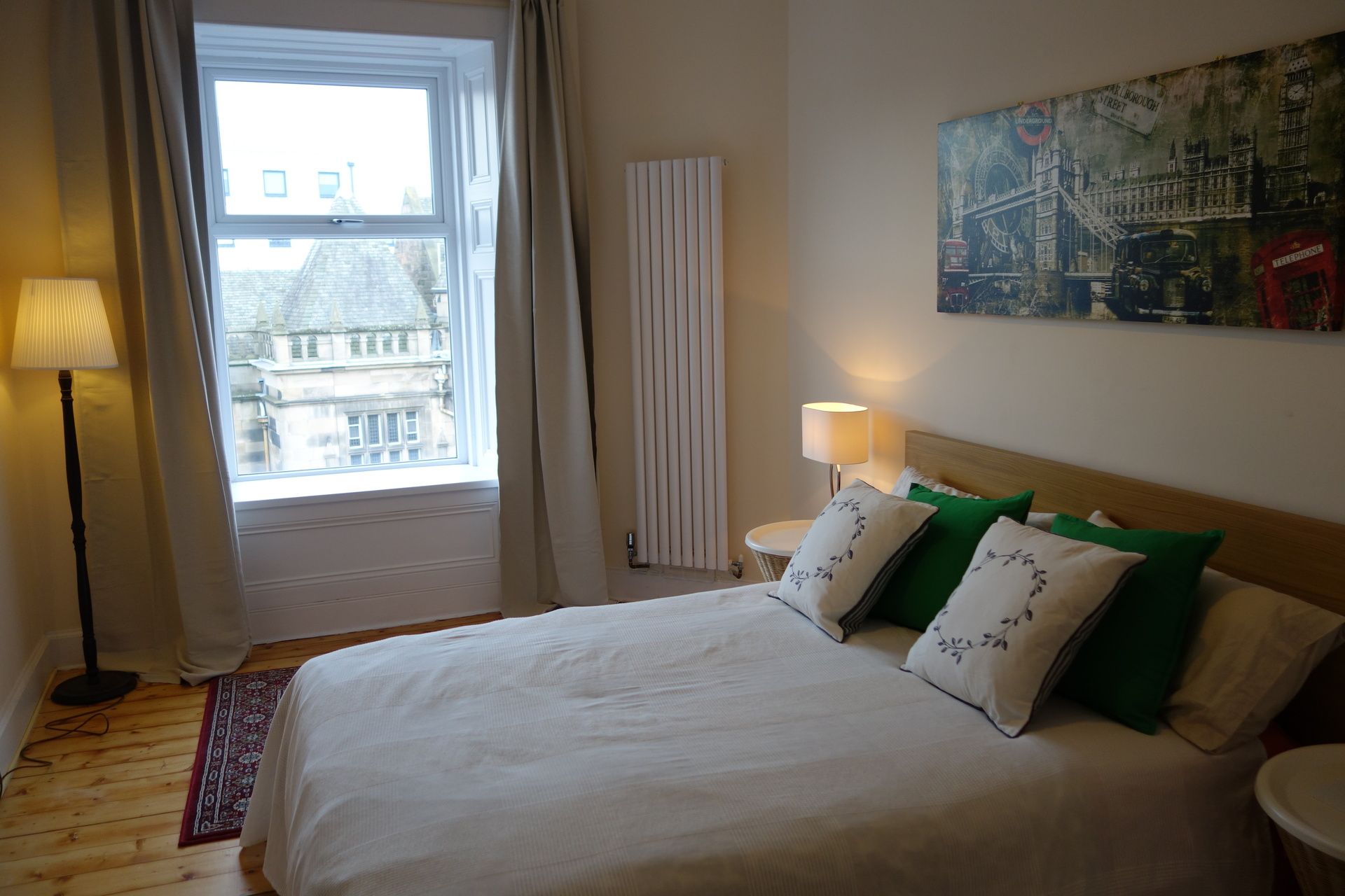 A hotel room with a bed, a window with curtains, a radiator, a lamp, and a painting on the wall.