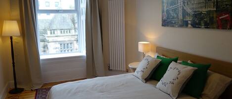 A hotel room with a bed, a window with curtains, a radiator, a lamp, and a painting on the wall.