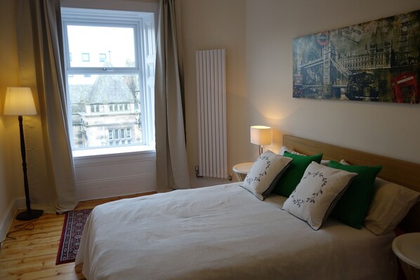 A hotel room with a bed, a window with curtains, a radiator, a lamp, and a painting on the wall.