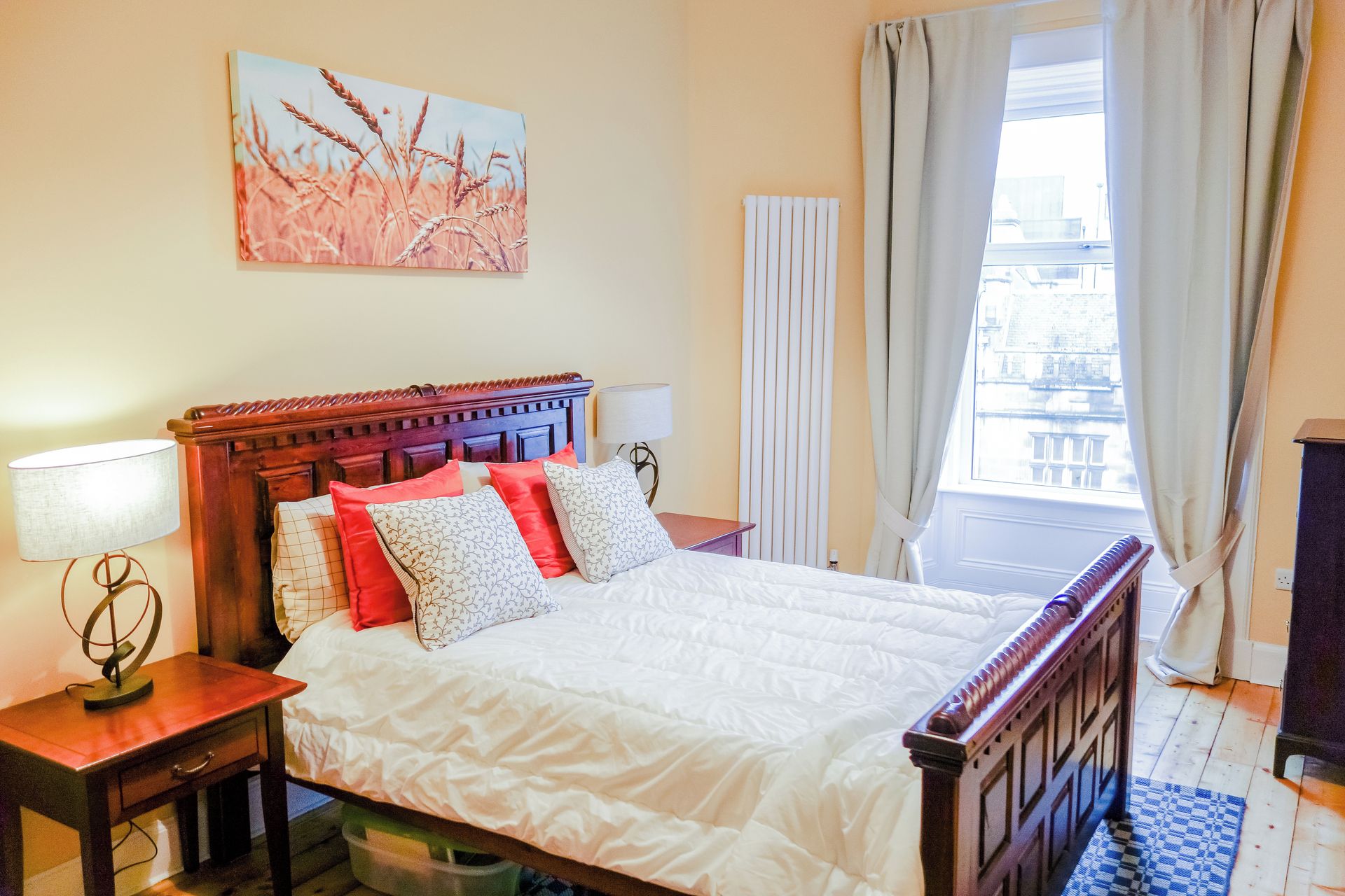 A hotel room with a bed, a nightstand, a lamp, a radiator, a window with curtains, and a city view.