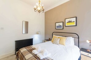 Un dormitorio con cama, radiador, lámpara de araña, lámpara de noche y dos cuadros en la pared.