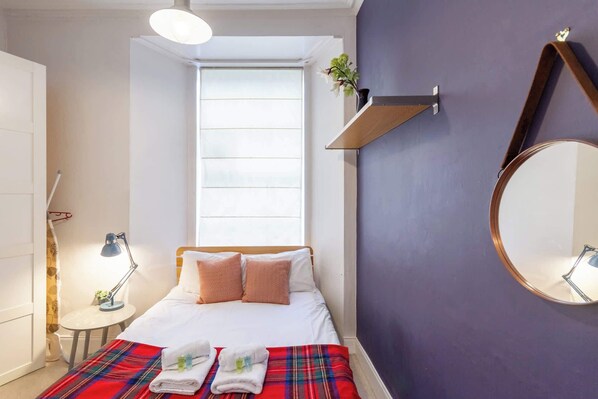 A bedroom with a bed, a window, a mirror, a bedside table, and a lamp.