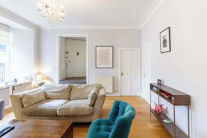 A living room with a sofa, a teal armchair, a wooden coffee table, a radiator, and a framed picture on the wall.