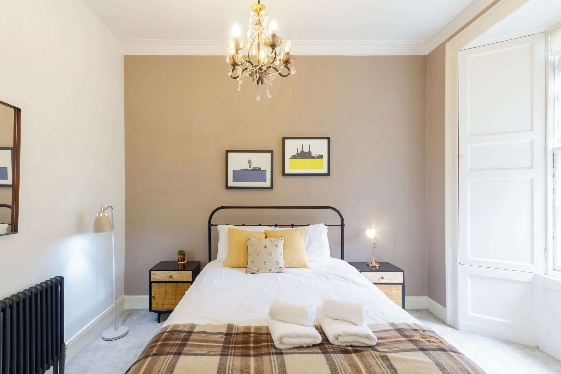 A bedroom with a bed, bedside tables, a radiator, a chandelier, and two framed pictures on the wall.