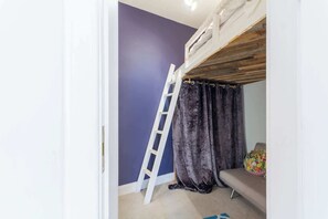 Una habitación con una litera, una escalera, un cortinaje oscuro y un cojín de colores sobre un sofá.