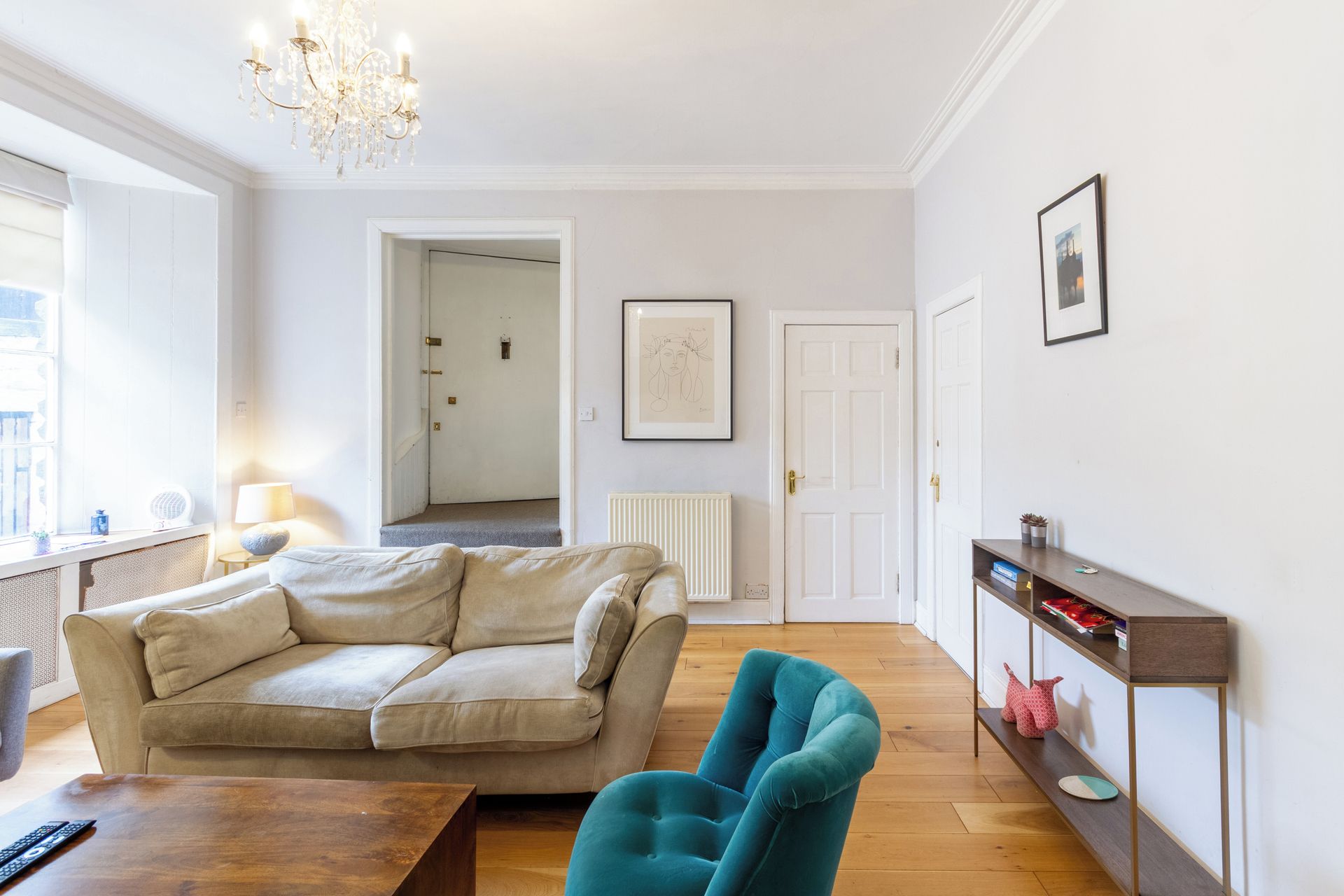 Ein Wohnzimmer mit einem beigefarbenen Sofa, einem türkisfarbenen Sessel, einem Holztisch und einer Heizung. Ein Flur führt in einen weiteren Raum, eine Tür und ein gerahmtes Bild sind an der Wand zu sehen.