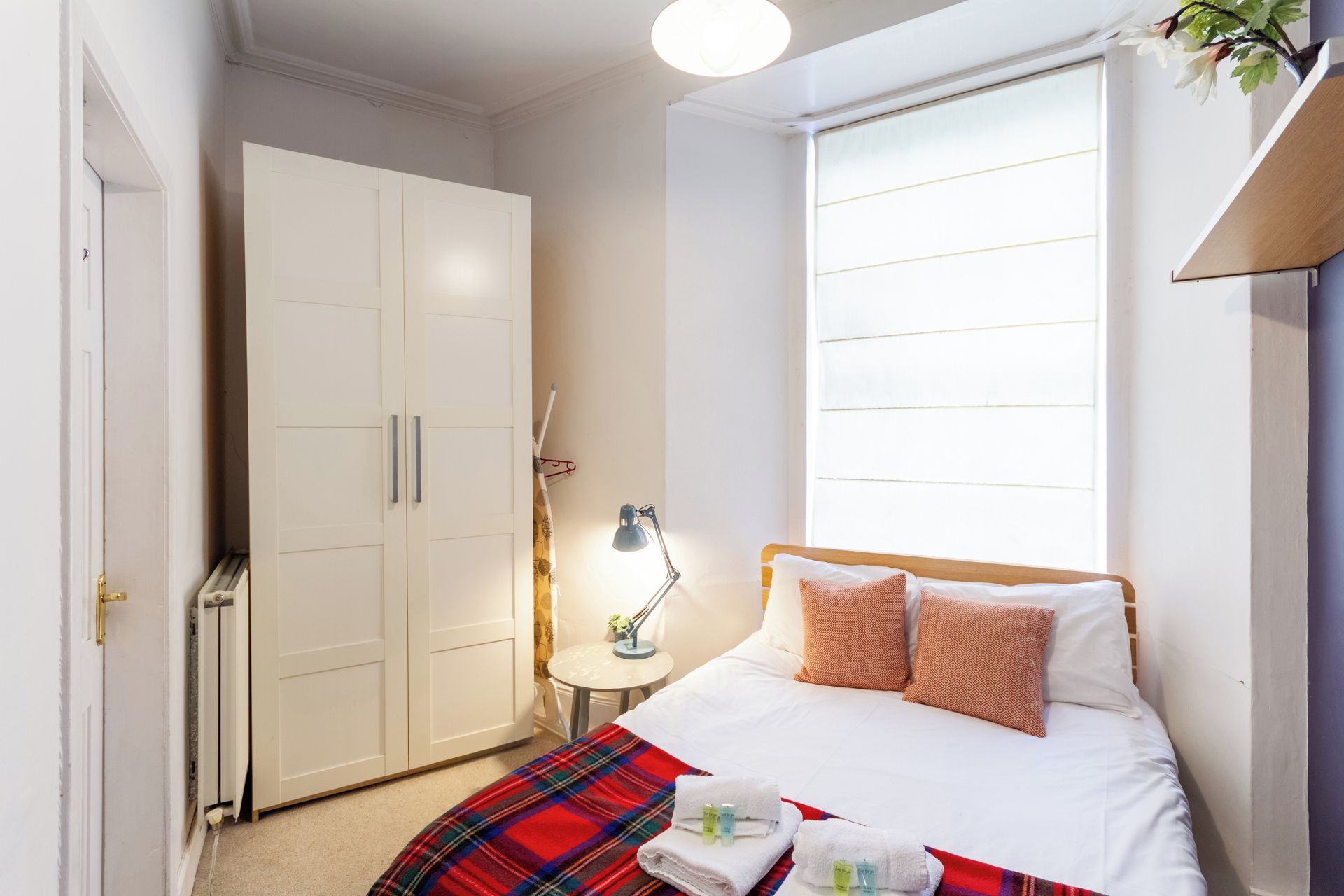 A bedroom with a bed, a nightstand, a lamp, a wardrobe, and a window.